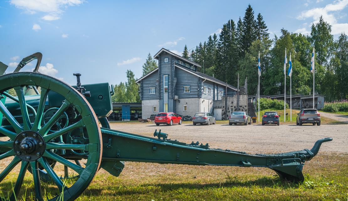 Kuva Taistelijan Talolta, joka esitt&auml;&auml; etualalla suurta, tummanvihre&auml;&auml;, vanhaa tykki&auml; tai haupitsia. Tykin suuri, puinen py&ouml;r&auml; n&auml;kyy etualalla vasemmalla. Taustalla on moderni, tummansininen tai harmaa museorakennus, jonka edess&auml; on hiekkaparkkipaikka ja joitakin autoja. Rakennuksen edess&auml; nousee useita lipputankoja, joissa liehuvat Suomen liput. Taivas on sininen ja pilvinen.