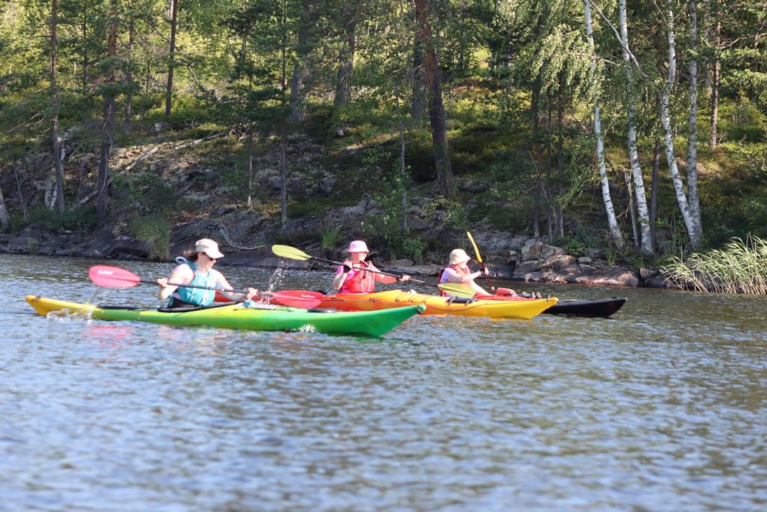 Kolme retkeilij&auml;&auml; meloo v&auml;rikk&auml;ill&auml; kajakeilla tyynell&auml; j&auml;rvell&auml; l&auml;hell&auml; kallioista ja mets&auml;ist&auml; rantaa.