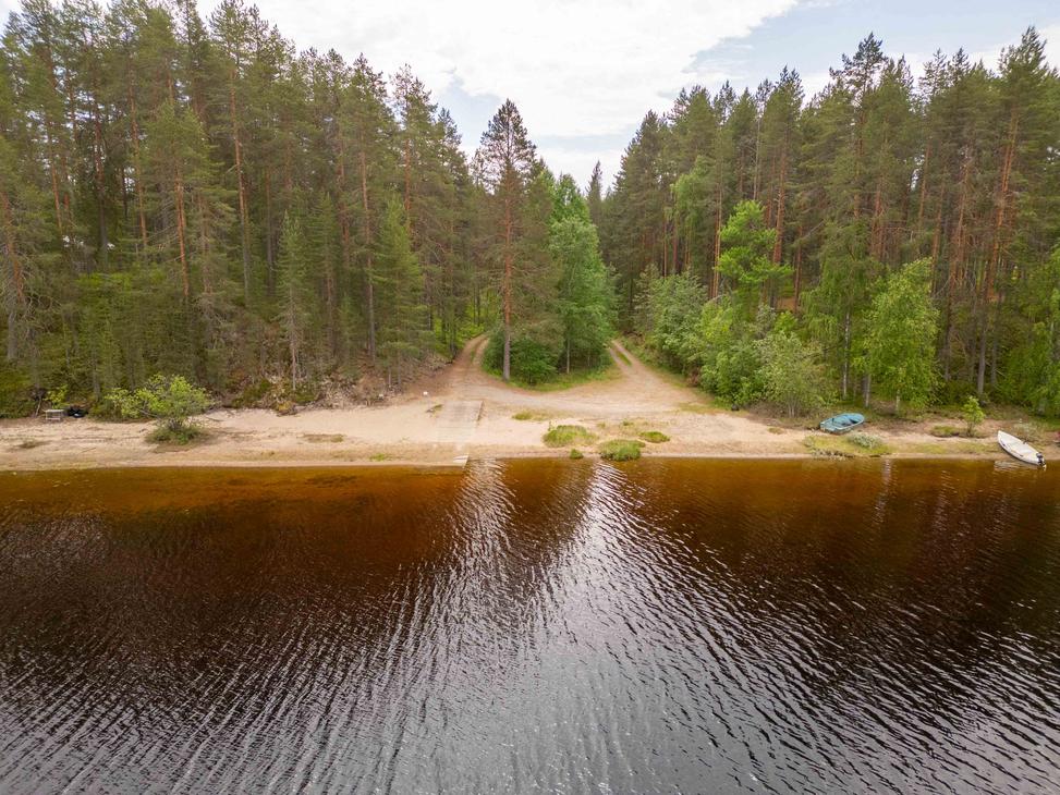 Ilmakuva järven hiekkarannasta ja veneenlaskupaikasta. Rantaan johtaa kaksi hiekkatietä tiheän mäntymetsän läpi, ja rantavedessä kimmeltää tumma vesi.