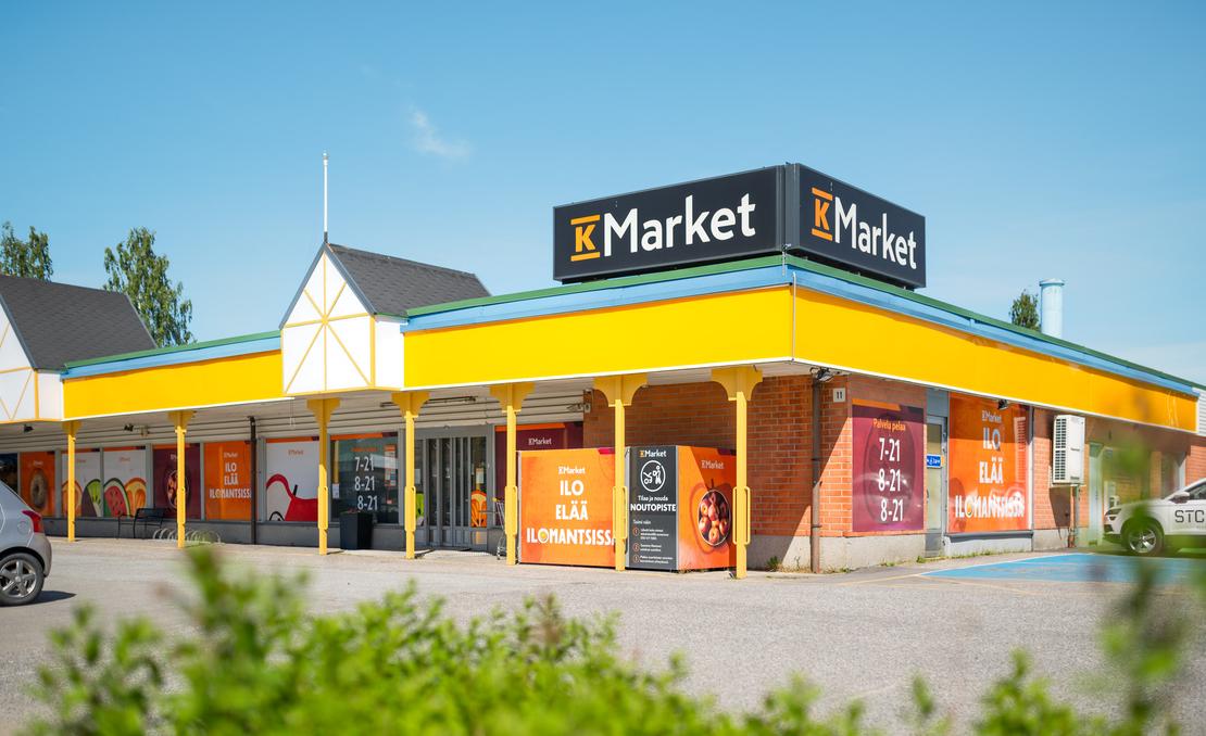 K-Market Ilomantsi -ruokakaupan julkisivu ja sis&auml;&auml;nk&auml;ynti aurinkoisena p&auml;iv&auml;n&auml; sinisen taivaan alla. Tiilirakennuksen katolla on suuret mustat K-Market-kyltit, ja katon reunaa kiert&auml;&auml; keltainen lippa. Ikkunoissa ja seiniss&auml; on oransseja opasteita, joissa lukee &rdquo;ILO EL&Auml;&Auml; ILOMANTSISSA&rdquo; sek&auml; kaupan aukioloajat. Sis&auml;&auml;nk&auml;ynnin l&auml;hell&auml; on noutopistekaappi, ja etualalla kasvaa vihre&auml;&auml; pensaikkoa.