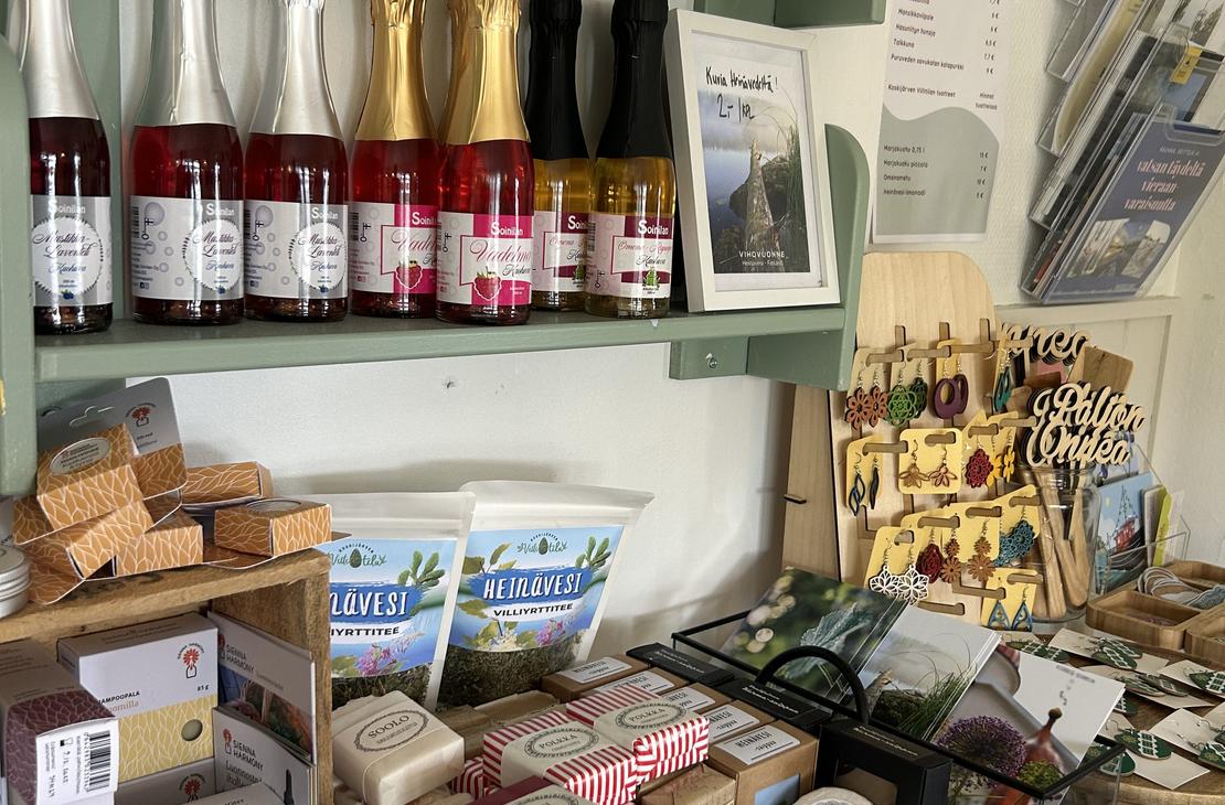 Lähikuva Satamakahvila-ravintola Kermacotan myymälähyllystä, jossa on tarjolla erilaisia paikallisia tuotteita ja matkamuistoja. Hyllyillä näkyy muun muassa pieniä kuohuviinipulloja, hilloja, paikallisia elintarvikkeita sekä käsitöitä. Tuotteet on aseteltu siististi puisiin ja vaaleanvihreisiin hyllykköihin.