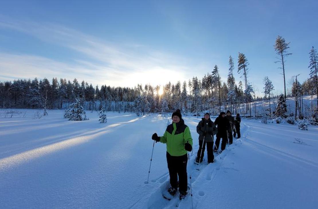 Ryhmä lumihiihdon tai lumikenkäilyn harrastajia hiihtää tai kävelee jonossa syvän, lumisen aukean yli kirkkaana talvipäivänä. Etualalla vasemmalla on yksi osallistuja kirkkaan limevihreässä takissa katsoen kameraan. Taustalla näkyy puiden ääriviivoja, jotka ovat lumen peitossa, ja aurinko paistaa matalalta horisontista luoden pitkiä varjoja.