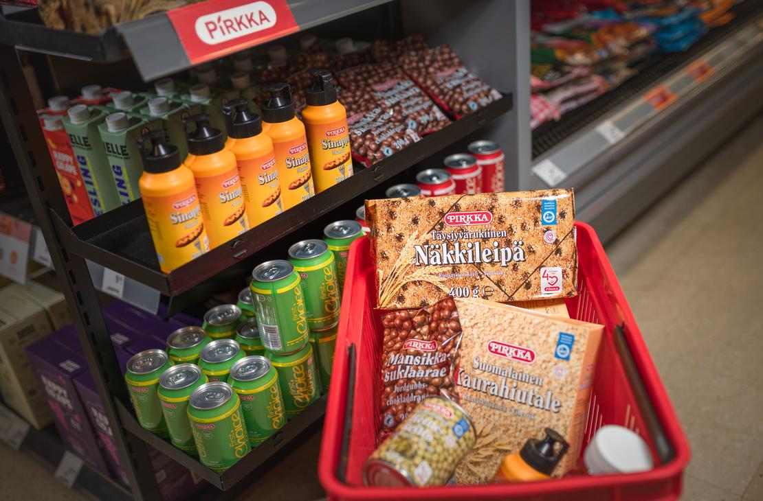 Punainen ostoskori t&auml;ynn&auml; Pirkka-merkkisi&auml; elintarvikkeita K-Market Ilomantsissa. Korissa on muun muassa Pirkka T&auml;ysjyv&auml;rukiinen N&auml;kkileip&auml;, Suomalainen Kaurahiutale ja Mansikkasuklaarae-pussi. Korin vieress&auml; on myym&auml;l&auml;n hylly, jossa on esill&auml; Pirkka-sinappeja ja vihreit&auml; juomat&ouml;lkkej&auml; punaisen Pirkka-kyltin alla.