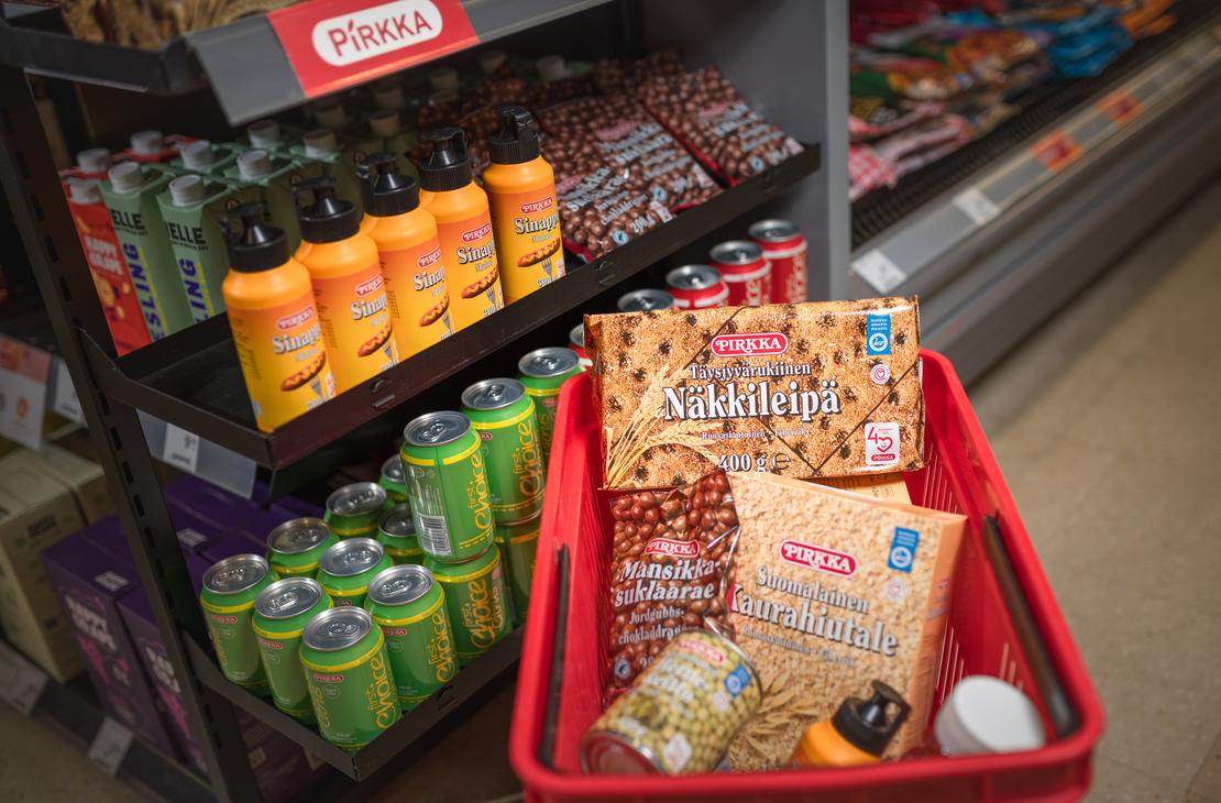 Punainen ostoskori täynnä Pirkka-merkkisiä elintarvikkeita K-Market Ilomantsissa. Korissa on muun muassa Pirkka Täysjyvärukiinen Näkkileipä, Suomalainen Kaurahiutale ja Mansikkasuklaarae-pussi. Korin vieressä on myymälän hylly, jossa on esillä Pirkka-sinappeja ja vihreitä juomatölkkejä punaisen Pirkka-kyltin alla.