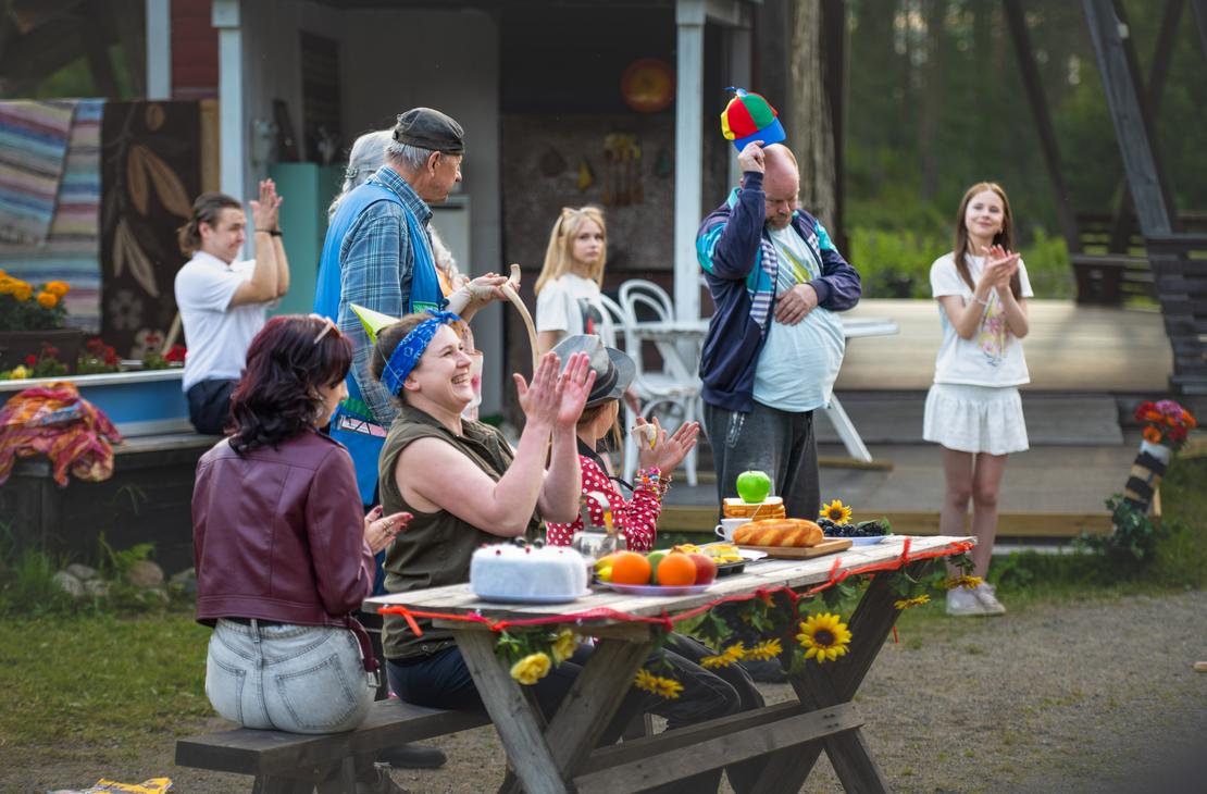 Möhkön kesäteatterin Peitetarina-näytelmän kohtaus, jossa useita näyttelijöitä on kokoontunut ulkonäyttämöllä herkuilla ja auringonkukilla koristellun pitkän puupöydän ääreen. Vasemmalla etualalla sinipäähineinen nainen nauraa ja taputtaa käsiään viininpunaiseen nahkatakkiin pukeutuneen naisen vieressä. Taustalla mies kohottaa värikästä potkurilippistään muiden näyttelijöiden taputtaessa ja seuratessa tilannetta.