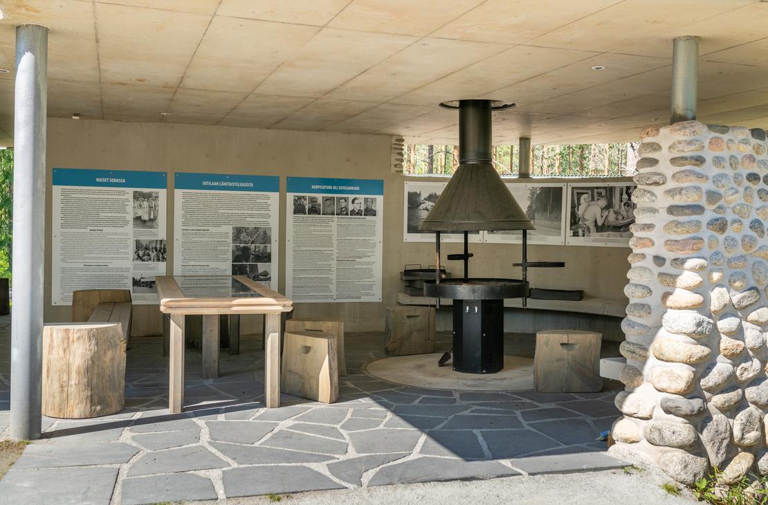 Sisäkuva Öykkösenvaaran taistelualueen taukopaikasta/museorakennuksesta. Kuvassa näkyy moderni, kivetty lattia, jolla on grillauspaikka tai takka suuren mustan liesituulettimen alla. Seinällä on kolme infotaulua teksteineen ja mustavalkokuvineen. Edessä on yksinkertainen puinen pöytä ja istuimina toimivia puunrunkoja. Oikealla näkyy luonnonkivillä verhoiltu pilarin osa. Tila on valoisa ja katettu.