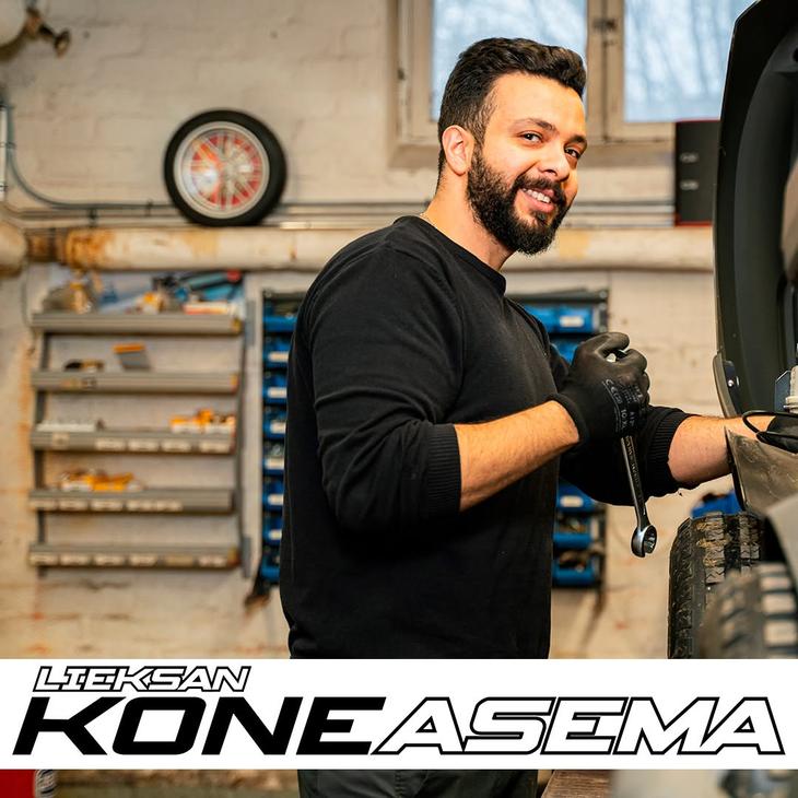 Parrakas mekaanikko hymyilee kameralle kiintoavain kädessään korjaamolla ajoneuvon äärellä. Kuvan alalaidassa on Lieksan Koneasema -tekstilogo.