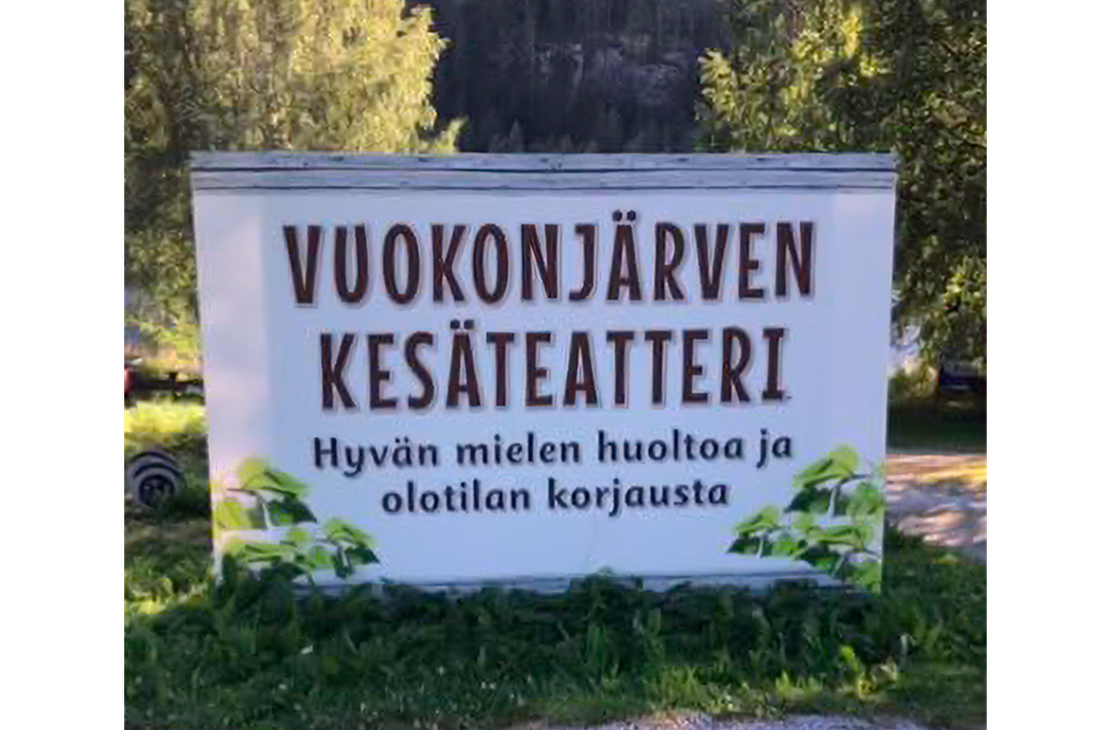Kuva esitt&auml;&auml; Vuokonj&auml;rven Kes&auml;teatterin opaskyltti&auml; ulkona luonnon helmassa. Kyltti on suorakulmainen ja valkoinen. Siin&auml; lukee tummanruskealla, isolla fontilla kaksi rivi&auml;: "VUOKONJ&Auml;RVEN" ja "KES&Auml;TEATTERI". Sen alapuolella on pienemm&auml;ll&auml;, mustalla tekstill&auml; tunnuslause "Hyv&auml;n mielen huoltoa ja olotilan korjausta". Kyltti on sijoitettu nurmikolle ja soraan, ja sit&auml; kehyst&auml;&auml; ala- ja sivuosissa vihre&auml; lehtikuviointi. Taustalla n&auml;kyy rehev&auml;&auml; mets&auml;&auml;.