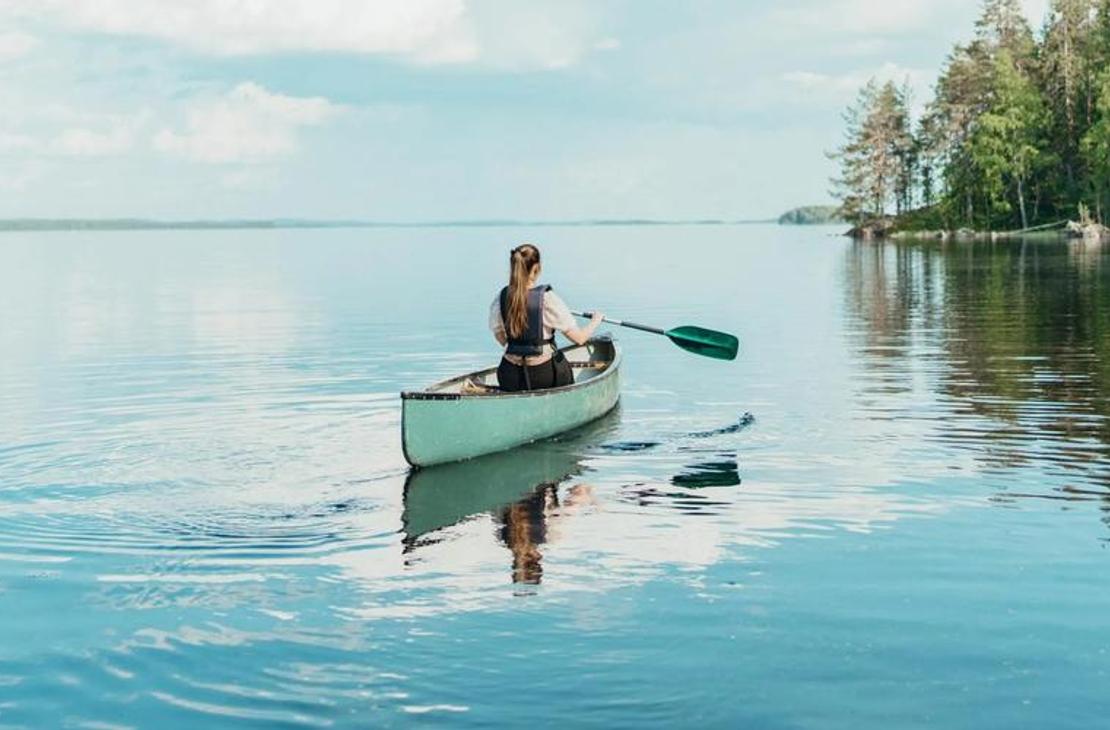 Kuva esittää naista melomassa yksinään vaaleanvihreällä kanootilla tyynellä järvellä aurinkoisena päivänä, liittyen Äksyt Ämmät -yrityksen melontapalveluihin. Nainen istuu kanootin perässä, meloo vihreällä melalla, ja hänen heijastuksensa näkyy tyynen sinisessä vedessä. Kanootin edessä on tyhjä tila. Taustalla näkyy metsäinen rantaviiva ja horisontissa aukea vesialue.
