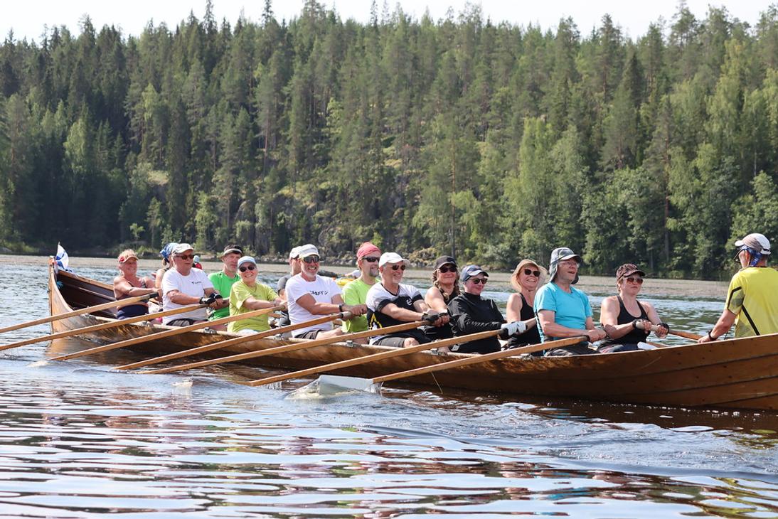 Pitkä puinen kirkkovene täynnä soutajia lipuu tyynellä järvellä. Taustalla näkyy jyrkkä metsäinen rinnerynnäs.