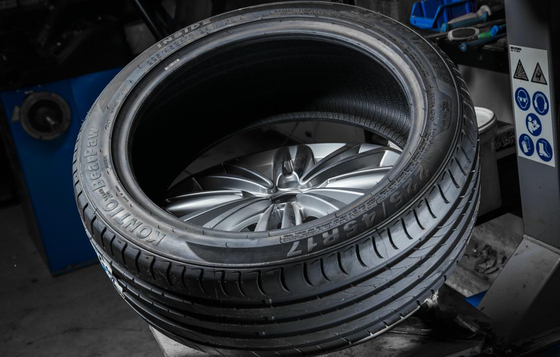 Lähikuva uudesta, matalaprofiilisesta renkaasta, joka on asennettavana kiiltävälle alumiinivanteelle. Renkaan pintakuvio on selkeä ja se on asetettu kaltevassa asennossa todennäköisesti rengaskoneen päällä. Taustalla näkyy epätarkkana sinisiä ja harmaita osia rengashuoltovälineistöä.