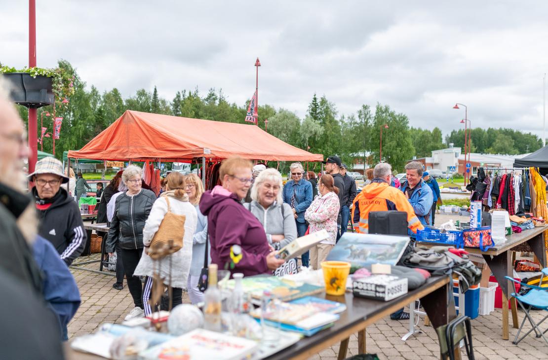 Ihmisiä tutkimassa tavaroita Ilomantsin ulkoilmatorin myyntipöydillä pilvisenä päivänä. Etualalla on pöytä täynnä pientavaraa, kuten kirjoja ja laatikoita, ja taustalla näkyy suuri oranssi myyntiteltta sekä vaaterekkejä.