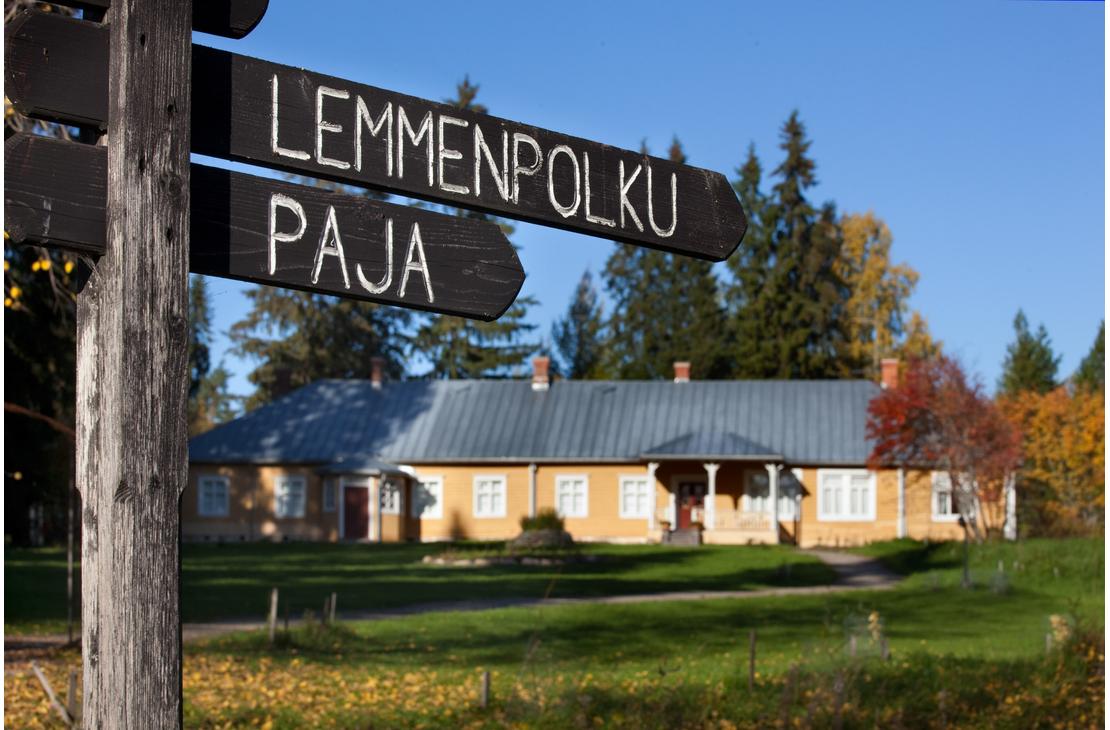 Musta puinen opasteviitta, jossa on valkoisella tekstit "LEMMENPOLKU" ja "PAJA". Taustalla näkyy epätarkkana Möhkön ruukkimuseon keltainen Pytinki-rakennus ja aurinkoinen nurmipiha.