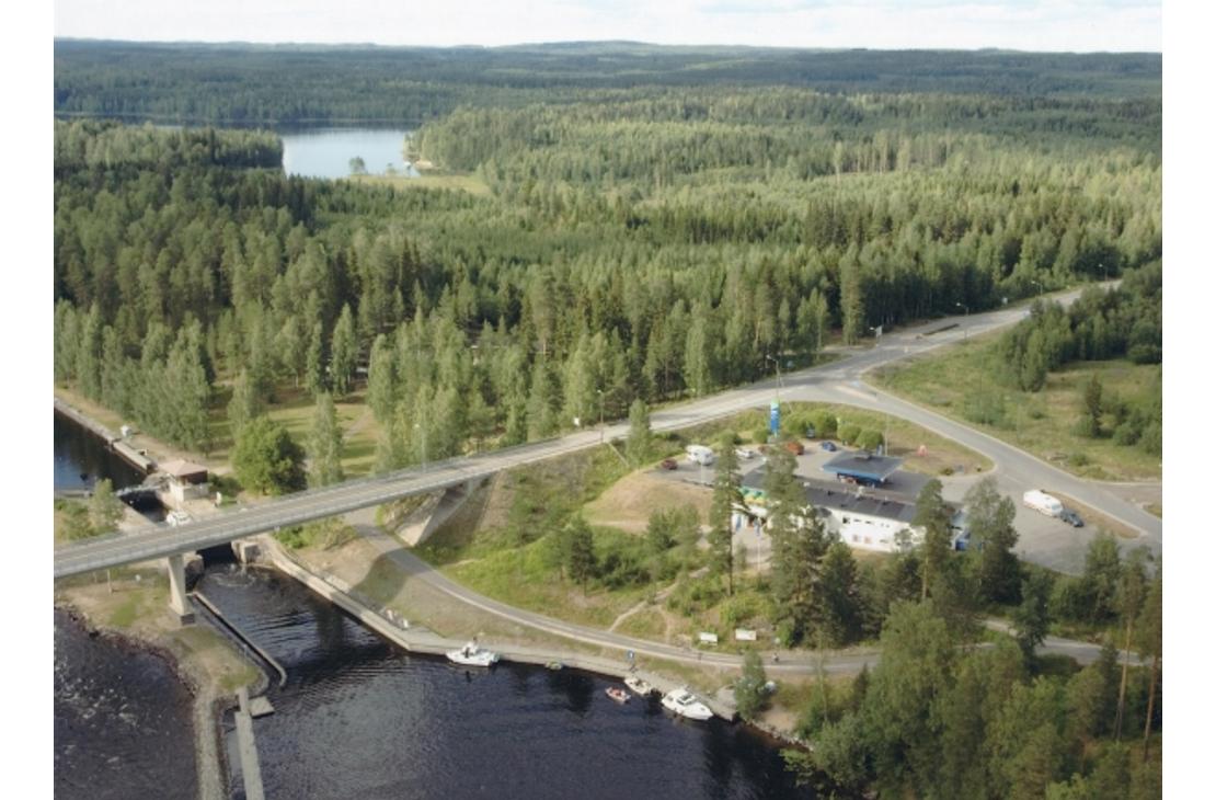 Ilmakuva Karvion kanavalta, jossa valtatie 23 ylittää kanavan siltaa pitkin. Kuvassa näkyy etualalla kanavan sulkuportteja ja laitureihin kiinnitettyjä veneitä. Tien vieressä sijaitsee Neste Karvionkanava -huoltoasema rakennuksineen ja parkkialueineen. Taustalla avautuu laaja ja vehreä metsämaisema sekä kaukana siintävä vesistö.