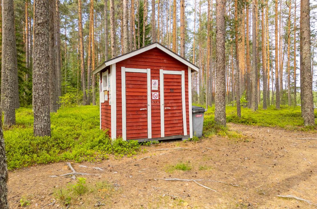 Punainen valkonurkkainen kuivakäymälä rakennus mäntymetsän keskellä. Rakennuksen vieressä on harmaa roska-astia ja maasto on käpystä kangasmaata.