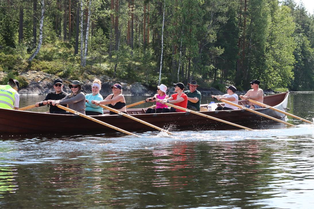 Sivukuva kirkkoveneest&auml;, jossa kahdeksan retkeilij&auml;&auml; soutaa tahdissa aurinkoisella j&auml;rvell&auml; mets&auml;isen rannan tuntumassa.
