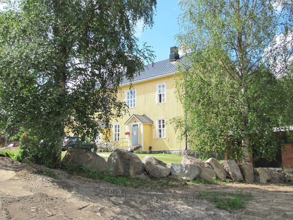 Katri Vala -kulttuurikeskuksen keltainen, kaksikerroksinen puurakennus aurinkoisena kesäpäivänä vihreiden koivujen ympäröimänä. Etualalla on hiekkamaata ja suurista luonnonkivistä koostuva matala muuri. Rakennuksessa on tumma harjakatto, valkoiset ikkunanpuitteet ja pieni katettu kuisti harmaan ulko-oven päällä.