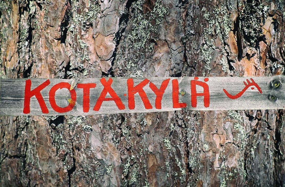 Lähikuva vanhan puun (todennäköisesti männyn) karkeasta, halkeilevasta kuoresta. Puunrunkoa vasten on kiinnitetty vaakasuora, vaaleampi puulauta, johon on maalattu kirkkaan punaisella tekstillä sana "KOTAKYLÄ". Tekstin oikeassa reunassa on pieni, punainen, tyylitelty kuvio, joka muistuttaa sarvipäätä tai eläintä.