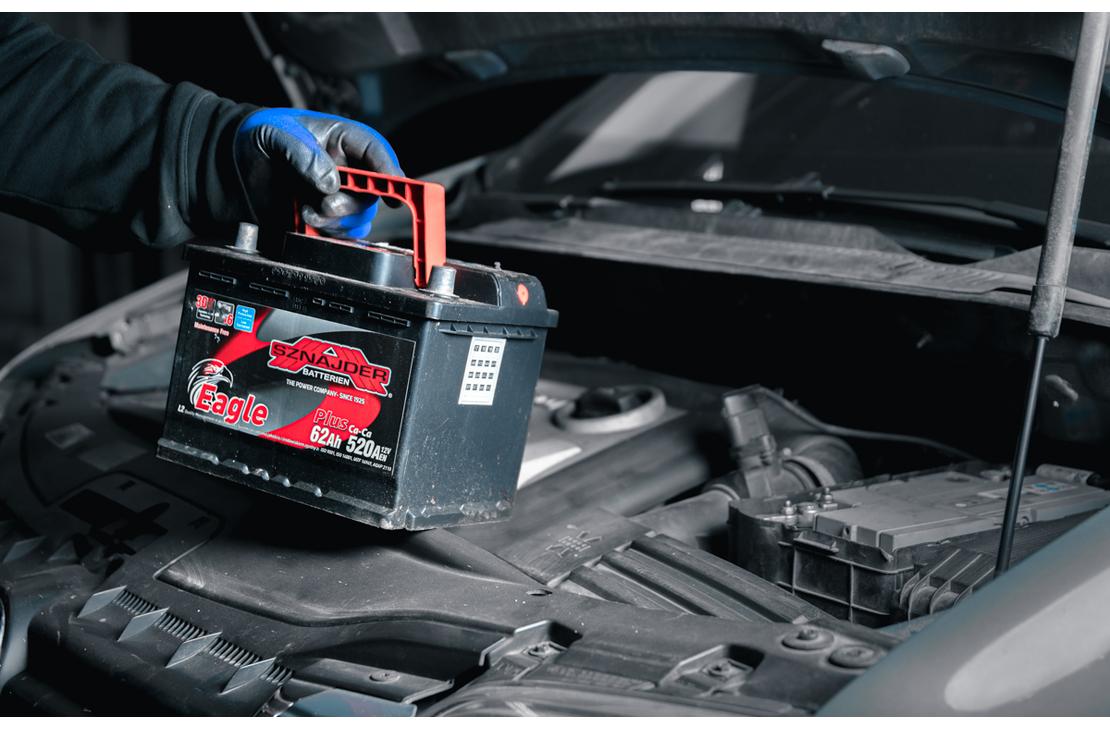 Lähikuva auton akun vaihdosta tai asennuksesta. Kuvassa nähdään käsi sinisessä hansikkaassa nostamassa tai laskemassa uutta, mustaa auton akkua, jossa on punainen kantokahva ja etiketti, harmaasävyisessä auton moottoritilassa. Konepelti on auki.