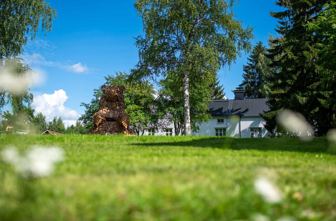 Laajakuva suuresta, puusta tehdystä karhupatsaasta, ILOKARHU -nimisestä veistoksesta Ilomantsista. Karhu on koottu useista pienemmistä puupaloista ja laudoista. Kuvassa näkyy selvästi karhun karkea pintakuvio. Taustalla on vihreää kasvillisuutta ja sininen taivas. Etualalla on muutama valkoinen kukka.