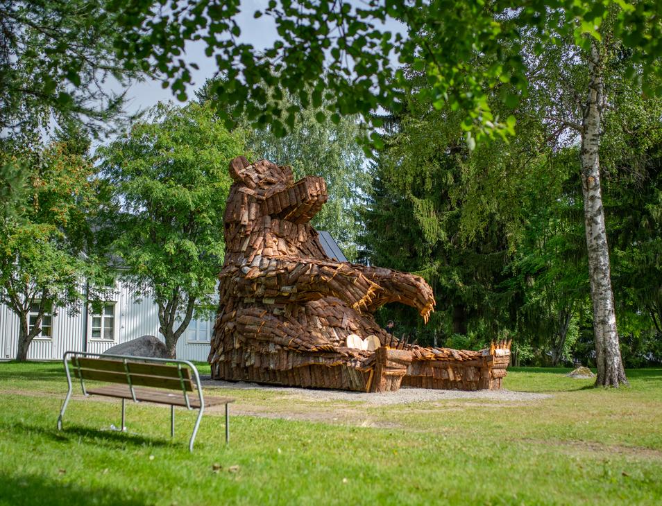 Lähikuva suuresta, puusta tehdystä karhupatsaasta, ILOKARHU -nimisestä veistoksesta Ilomantsista. Karhu on koottu useista pienemmistä puupaloista ja laudoista. Kuvassa näkyy selvästi karhun karkea pintakuvio. Taustalla on vihreää kasvillisuutta.