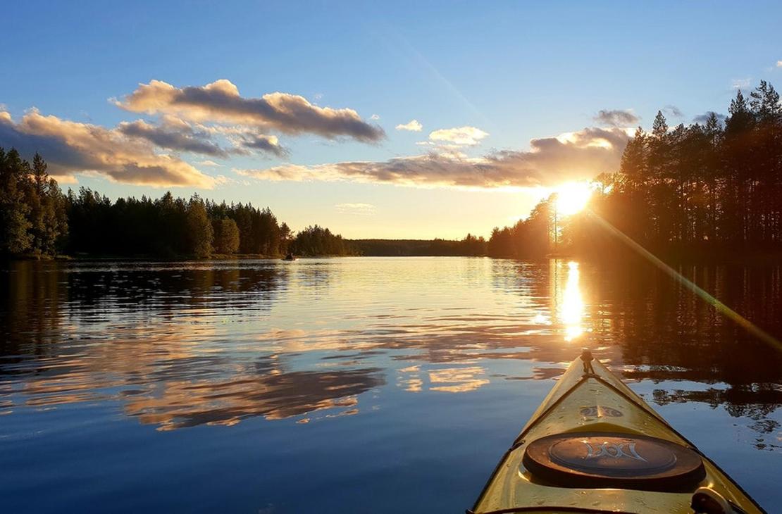 Kuva on otettu kajakista tai kanootista, ja se esitt&auml;&auml; tyynt&auml; j&auml;rve&auml; auringonlaskun aikaan. Kajakin keula (vaaleankeltainen) on etualalla. Aurinko laskee tai nousee suoraan edess&auml;, luoden kirkkaan heijastuksen veteen, joka on sinist&auml; ja heijastaa taivaan pilvi&auml;. J&auml;rve&auml; reunustaa tumma, tihe&auml; mets&auml; molemmin puolin. Kuva liittyy todenn&auml;k&ouml;isesti &Auml;ksyt &Auml;mm&auml;t -yrityksen melontapalveluihin.