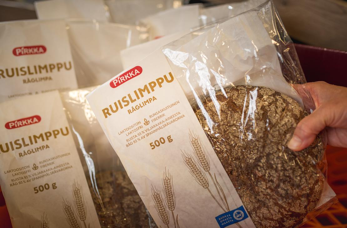 Käsi pitelee osittain läpinäkyvää Pirkka Ruislimppu -leipäpakkausta K-Market Ilomantsissa. 500 gramman pakkauksesta erottuu pyöreä ruisleipä sekä tekstit ”Pirkka Ruislimppu Råglimpa”. Taustalla on useita muita samanlaisia leipäpakkauksia rivissä.