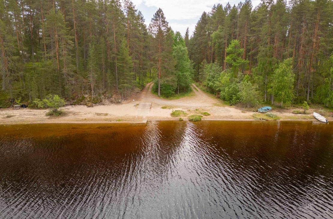 Ilmakuva järven hiekkarannasta ja veneenlaskupaikasta. Rantaan johtaa kaksi hiekkatietä tiheän mäntymetsän läpi, ja rantavedessä kimmeltää tumma vesi.