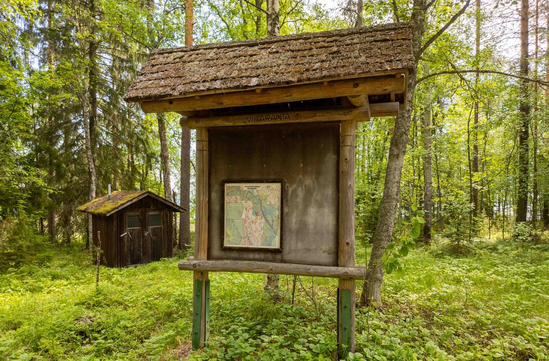 Puinen huopakattoinen opastetaulu metsän reunassa, jossa on suuri kartta ja teksti Viitaranta. Taustalla näkyy vanha tummapuinen kuivakäymälä.