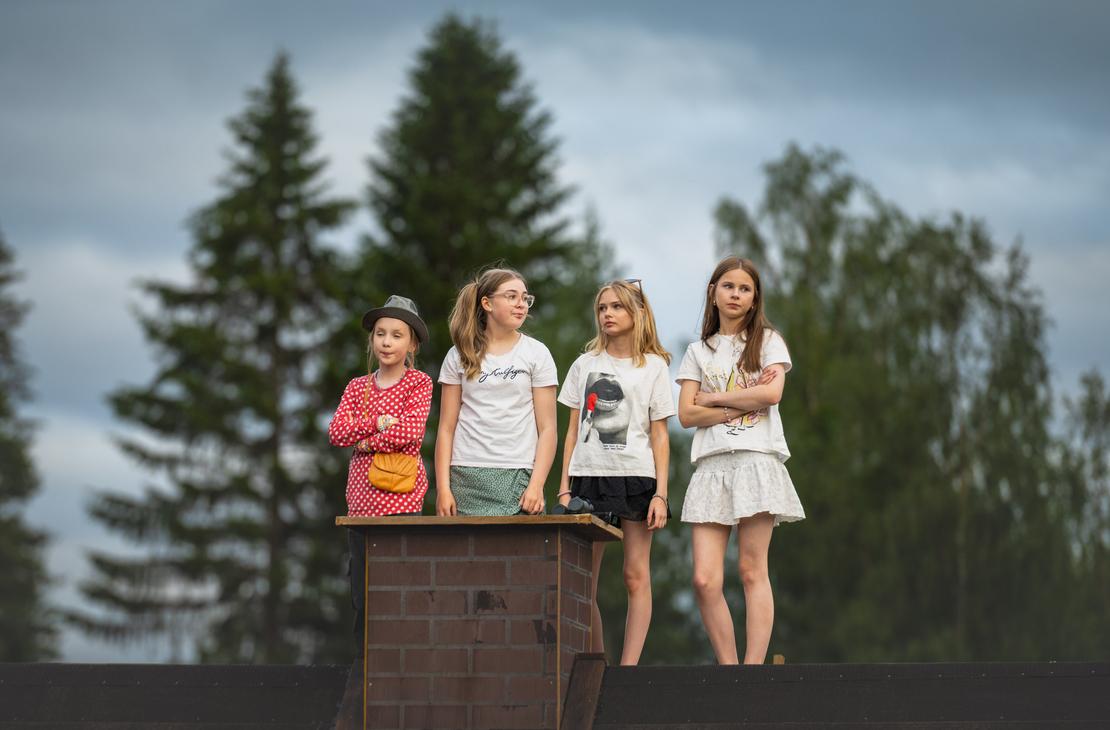 Neljä nuorta tyttöä seisoo vierekkäin tiilisavupiipun vieressä rakennuksen katolla Möhkön kesäteatterin Peitetarina-näytelmässä. Heillä on yllään kesäiset vaatteet, osa pitää käsiään puuskassa, ja he katsovat tarkkaavaisina sivuun taustallaan korkeat vihreät puut ja pilvinen taivas.