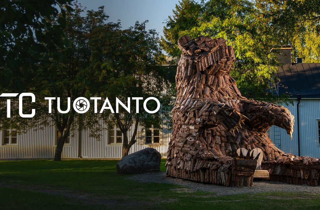 Kuva esittää pihanurmea, jolla istuu suuri, karhunmuotoinen veistos tai patsas, joka on tehty risuista, puunpaloista tai muusta tummasta materiaalista. Patsas on sijoitettu historiallisen, vaalean puurakennuksen (oikealla) ja valkoisen rakennuksen (vasemmalla) eteen. Kuvan päällä lukee valkoisella, läpikuultavalla tekstillä "STC TUOTANTO", ja T-kirjainten välissä on pieni pyöreä logo.