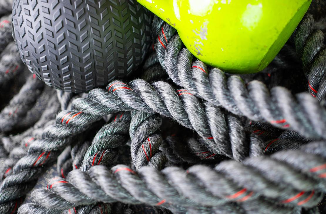 Lähikuva Kinesiocenter Kuntoilukeskuksen kuntosalilaitteista. Köysien ja painojen kasauma on etualalla: tummanharmaa harjoitusköysi (taisteluköysi), kirkkaan limevihreä kahvakuula ja tumma, kuvioitu painopallo (medicine ball). Kuva symboloi tehokasta ja monipuolista harjoittelua.