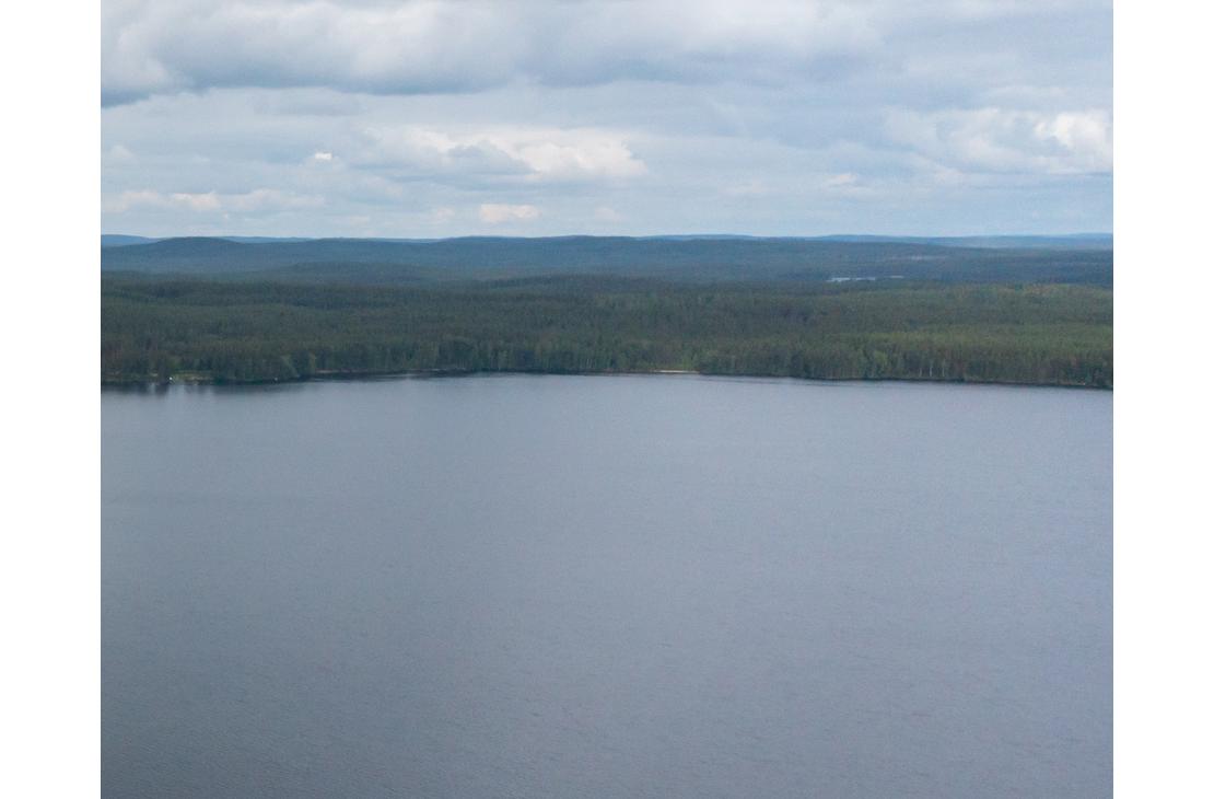 Laaja panoraamakuva, joka on otettu korkealta. Se esittää tyyntä, tummansinistä järven pintaa, joka täyttää suurimman osan kuvasta. Järven takana horisontissa näkyy tiheä, tummanvihreä metsä ja taustalla kaukaisia metsäisiä kukkuloita. Taivas on pilvinen ja vaaleanharmaa.