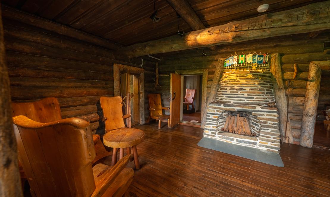 Sisäkuva Rajakenraalin majan oleskelutilasta. Huone on tunnelmallinen ja siinä on tummat hirsiseinät sekä puulattiat. Keskeisellä paikalla on suuri, luonnonkivistä muurattu takka. Vasemmalla näkyy kaksi nahkaverhoiltua tai puunväristä nojatuolia pienen sivupöydän ympärillä. Takan takana on oviaukkoja, jotka johtavat muihin tiloihin. Tila on valaistu lämpimällä, pehmeällä valolla.