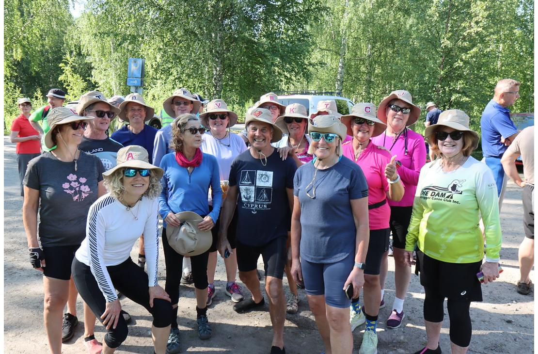 Ryhmä aurinkohattuihin pukeutuneita hymyileviä retkeilijöitä seisoo hiekkatiellä puiden katveessa. Useimpien hatuissa on yksittäinen kirjainmerkki.