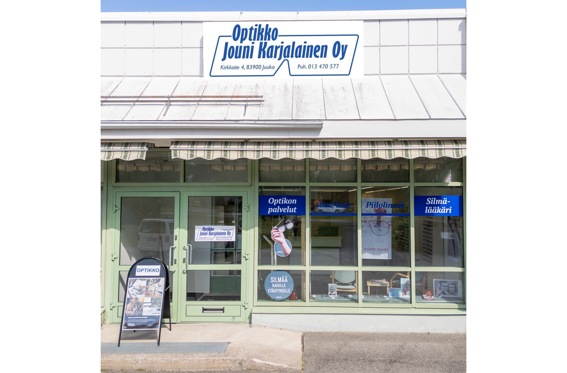 Kuva Optikko Jouni Karjalainen Oy -liikkeen ulkopuolesta aurinkoisena p&auml;iv&auml;n&auml;. Liikkeess&auml; on suuret ikkunat ja vaaleanvihre&auml; ovi. Ikkunoissa on mainosteippauksia, joissa mainitaan "Optikon palvelut", "Piilolinssit Kuno-Qvist" ja "Silm&auml;l&auml;&auml;k&auml;ri". Sis&auml;&auml;nk&auml;ynnin yl&auml;puolella on markiisi. Rakennuksen yl&auml;osassa on suuri, kaarevalla kehyksell&auml; varustettu kyltti, jossa lukee "Optikko Jouni Karjalainen Oy", sek&auml; osoite "Kirkkotie 4, 83900 Juuka" ja puhelinnumero "Puh. 013 470 577".