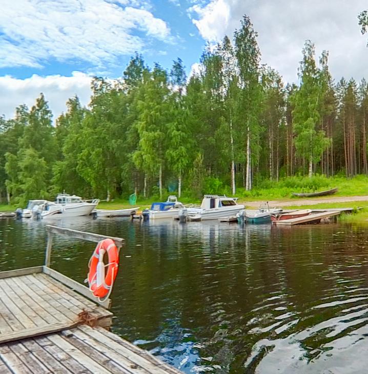 Kuva esittää pienvenesatamaa tai venelaituria aurinkoisena päivänä. Useita moottoriveneitä ja pienempiä soutuveneitä on kiinnitetty laituriin tumman veden äärellä. Etualalla vasemmalla on puinen laituri, jonka kulmassa on kiinni oranssi pelastusrengas. Taustalla näkyy tiheää metsää, jossa on sekä havupuita että vaaleita koivuja. Taivaalla on valkoisia pilviä.