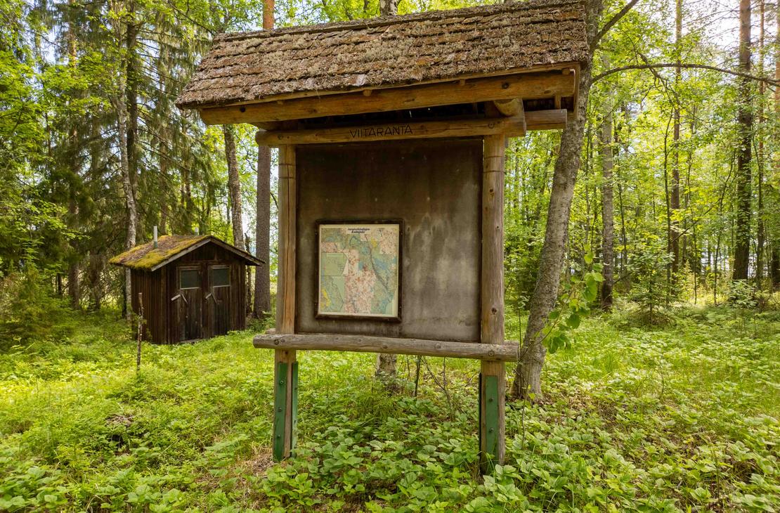 Puinen huopakattoinen opastetaulu metsän reunassa, jossa on suuri kartta ja teksti Viitaranta. Taustalla näkyy vanha tummapuinen kuivakäymälä.