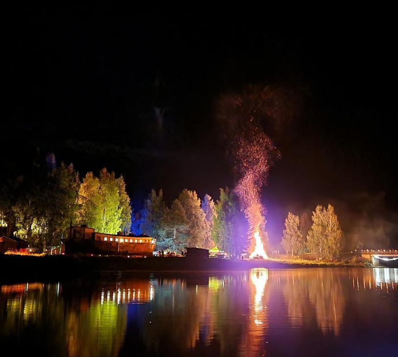 Yökuva Möhköstä vesistön äärellä järjestetystä tapahtumasta. Keskellä hehkuu suuri, korkea kokko tai roihuava nuotio, jonka liekit valaisevat taivaan. Kokko heijastuu kirkkaana tyyneen veteen etualalla. Rantaviivan puita on valaistu erivärisillä jouluvaloilla, luoden keltaisia, vihreitä ja violetteja sävyjä pimeään. Taustalla näkyy rakennuksen siluetti.