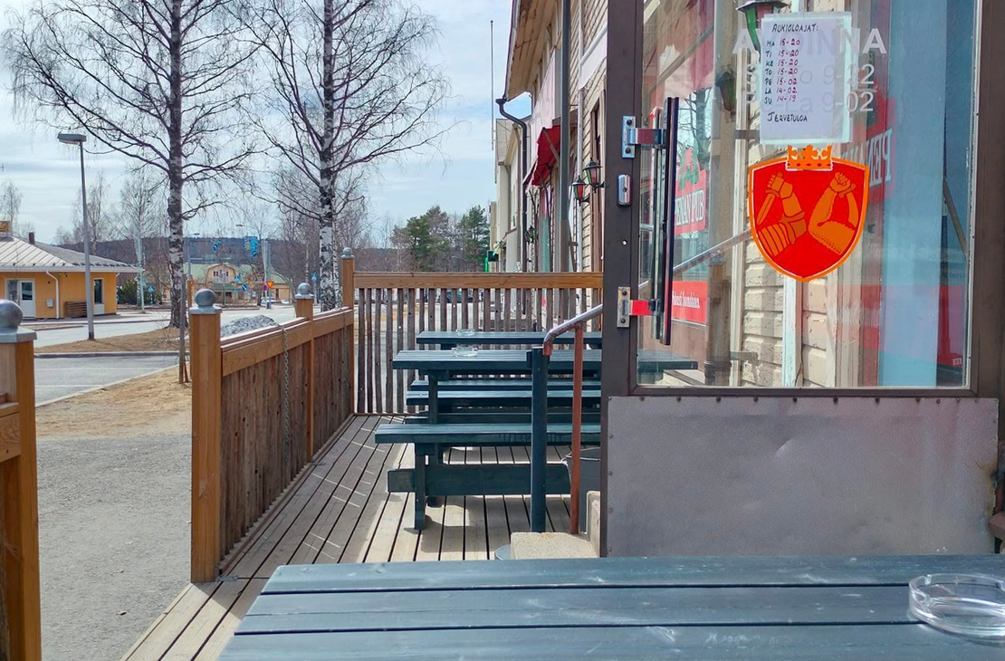 Kuva esittää Penan Pubin ulkoterassia aurinkoisena päivänä. Etualalla on puinen, tummansininen terassipöytä. Terassilla on puulattia ja tummansinisiä pöytä-penkkiyhdistelmiä. Terassin vieressä on rakennuksen lasiovi ja ikkuna, jossa on punainen, vihreä- ja kelta-oranssisävyinen "PENAN PUB" -kyltti näkyvissä. Taustalla näkyy katua, nurmikkoa ja koivuja, jotka ovat vielä lehdettömiä.