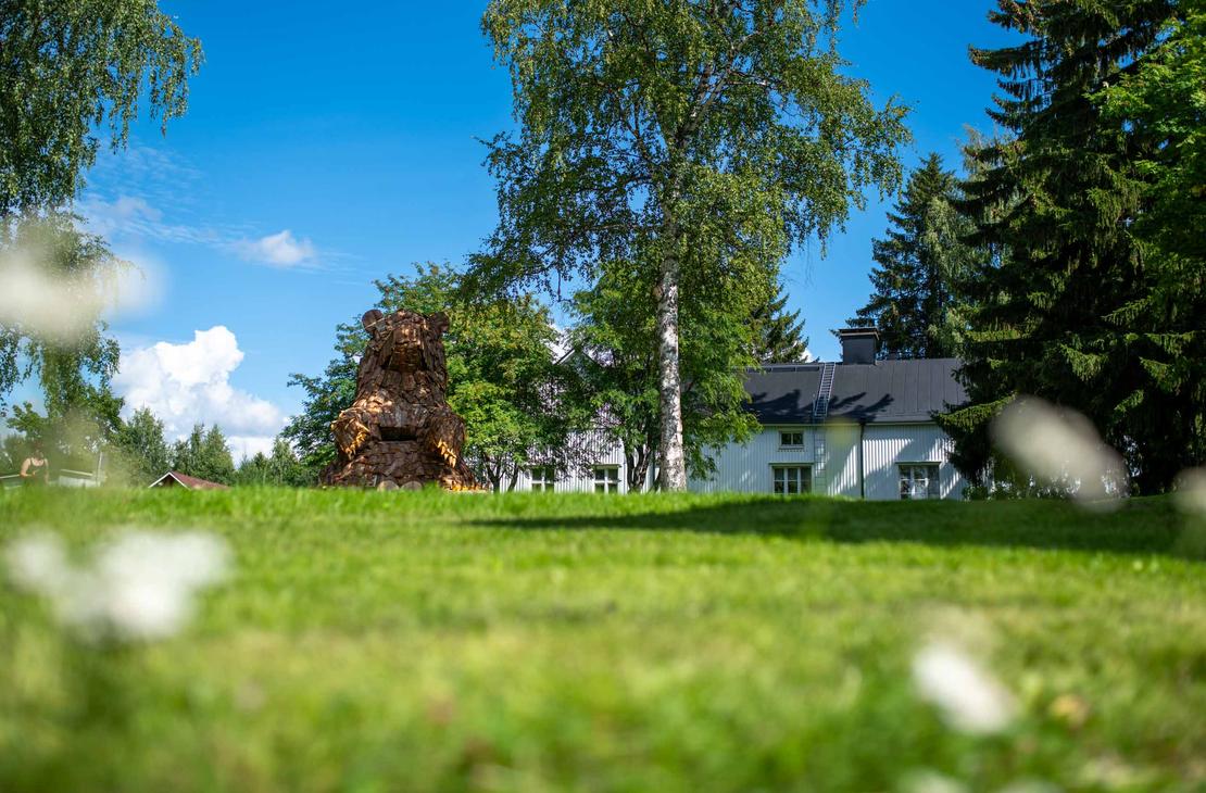 Laajakuva suuresta, puusta tehdystä karhupatsaasta, ILOKARHU -nimisestä veistoksesta Ilomantsista. Karhu on koottu useista pienemmistä puupaloista ja laudoista. Kuvassa näkyy selvästi karhun karkea pintakuvio. Taustalla on vihreää kasvillisuutta ja sininen taivas. Etualalla on muutama valkoinen kukka.