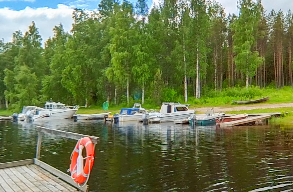 Kuva esittää pienvenesatamaa tai venelaituria aurinkoisena päivänä. Useita moottoriveneitä ja pienempiä soutuveneitä on kiinnitetty laituriin tumman veden äärellä. Etualalla vasemmalla on puinen laituri, jonka kulmassa on kiinni oranssi pelastusrengas. Taustalla näkyy tiheää metsää, jossa on sekä havupuita että vaaleita koivuja. Taivaalla on valkoisia pilviä.