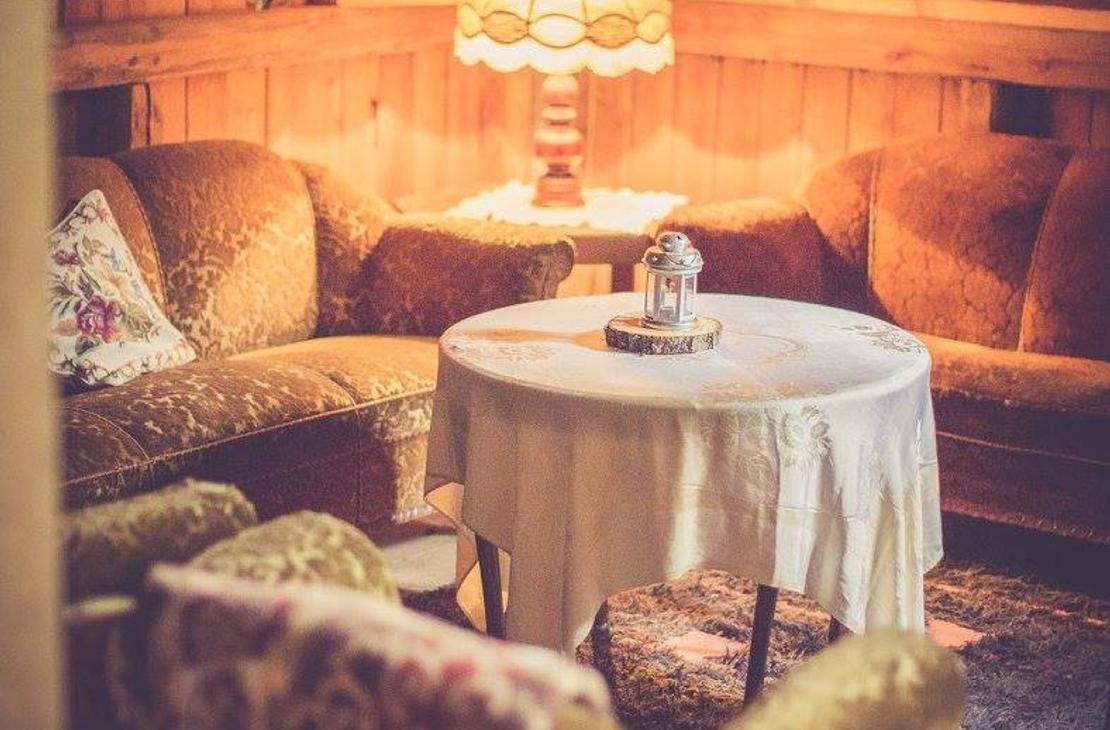 Kuva esitt&auml;&auml; Pappila Marjam&auml;en ullakolla tai parvella sijaitsevaa oleskelutilaa. Tila on sisustettu kodikkaasti retrotyylisill&auml;, sammaleenvihreill&auml; tai oranssinruskeilla sohvilla ja nojatuoleilla. Huoneessa on puupaneelisein&auml;t ja -katto. Keskell&auml; on py&ouml;re&auml; p&ouml;yt&auml; valkoisella p&ouml;yt&auml;liinalla, ja tila on valaistu l&auml;mpim&auml;sti vanhanaikaisella p&ouml;yt&auml;valaisimella. Taustalla n&auml;kyy kattorakenteita ja yl&auml;tasolle viev&auml;t tikkaat.