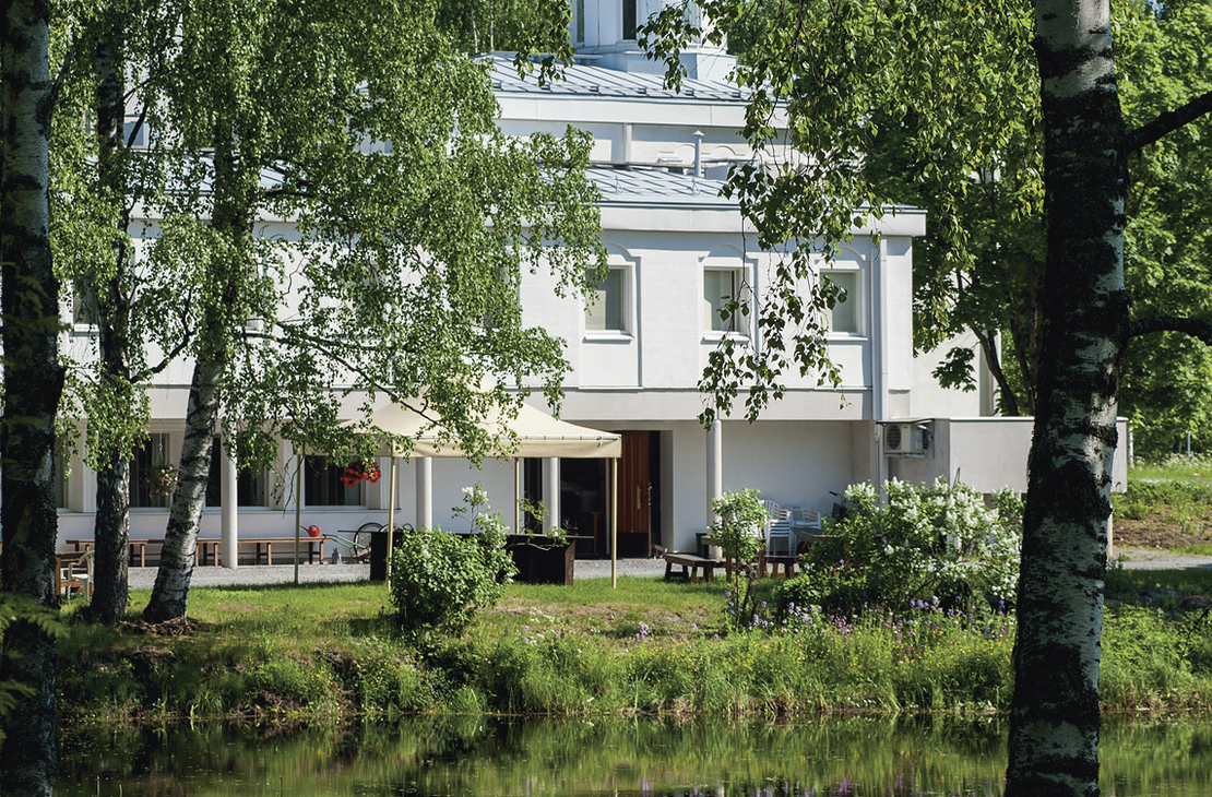 Lintulan luostarin valkoinen rakennus kuvattuna veden ääreltä kesäisenä päivänä. Etualalla on lampi, josta rakennus ja sitä kehystävät koivut heijastuvat.