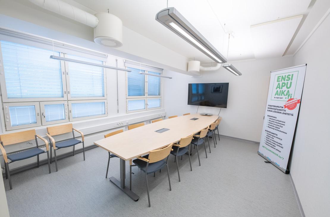 Kuva esittää Liikekulman Toimistohotellin isoa, valoisaa neuvottelutilaa tai koulutushuonetta. Huoneessa on vaalea lattia, ja keskellä on suuri, vaaleapuinen neuvottelupöytä, jonka ympärillä on mustia tuoleja. Pöydän päällä on liitäntöjä. Oikealla seinällä on suuri taulutelevisio ja pystyssä oleva roll-up-mainos. Vasemmalla on pitkä ikkunanauha, jossa on siniset sälekaihtimet, ja seinää vasten on muutama ylimääräinen vierastuoli.