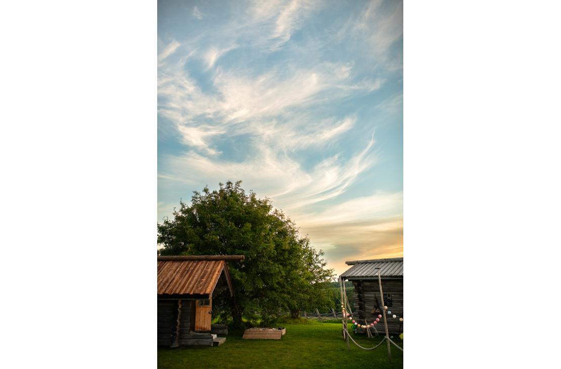 Ravintola Parppeinpirtti, Kaksi pientä hirsirakennusta ja suuri lehtipuu vihreällä nurmialueella. Yläpuolella on sininen taivas, jolla on valkoisia haituvapilviä.