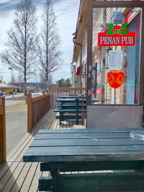 Kuva esittää Penan Pubin ulkoterassia aurinkoisena päivänä. Etualalla on puinen, tummansininen terassipöytä. Terassilla on puulattia ja tummansinisiä pöytä-penkkiyhdistelmiä. Terassin vieressä on rakennuksen lasiovi ja ikkuna, jossa on punainen, vihreä- ja kelta-oranssisävyinen "PENAN PUB" -kyltti näkyvissä. Taustalla näkyy katua, nurmikkoa ja koivuja, jotka ovat vielä lehdettömiä.