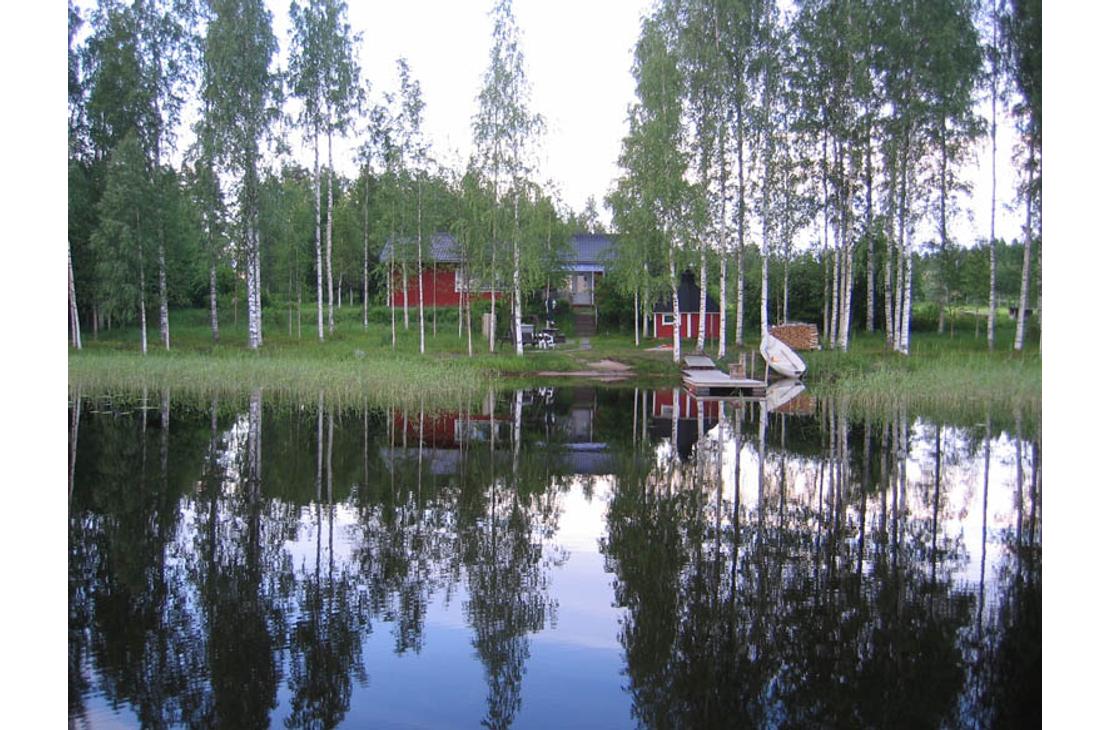 Kuva esittää Villa Blancan / LillaVilla Villan rantamaisemaa järveltä päin. Tyyni vesi etualalla heijastaa kirkkaasti taustalla olevia hoikkia koivuja ja muita puita. Puiden takaa erottuu punamultainen mökki tai sauna. Rannassa on pieni laituri ja mahdollisesti valkoinen vene tai kanootti. Maisema on rauhallinen ja kesäinen.