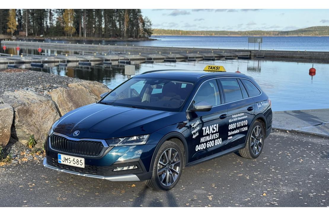 Kuva esittää tummansinistä, farmarimallista taksia (Skoda Octavia) pysäköitynä satama-alueella. Auton katolla on keltainen "TAKSI" -kyltti. Kyljessä on useita tekstejä, joista erottuvat selkeästi sanat "TAKSI HEINÄVESI 0400 600 800" valkoisella, sekä puhelinnumeroita. Auto on pysäköity sora-alueelle suuren kiven viereen. Taustalla näkyy tyyni järvimaisema ja venelaitureita.
