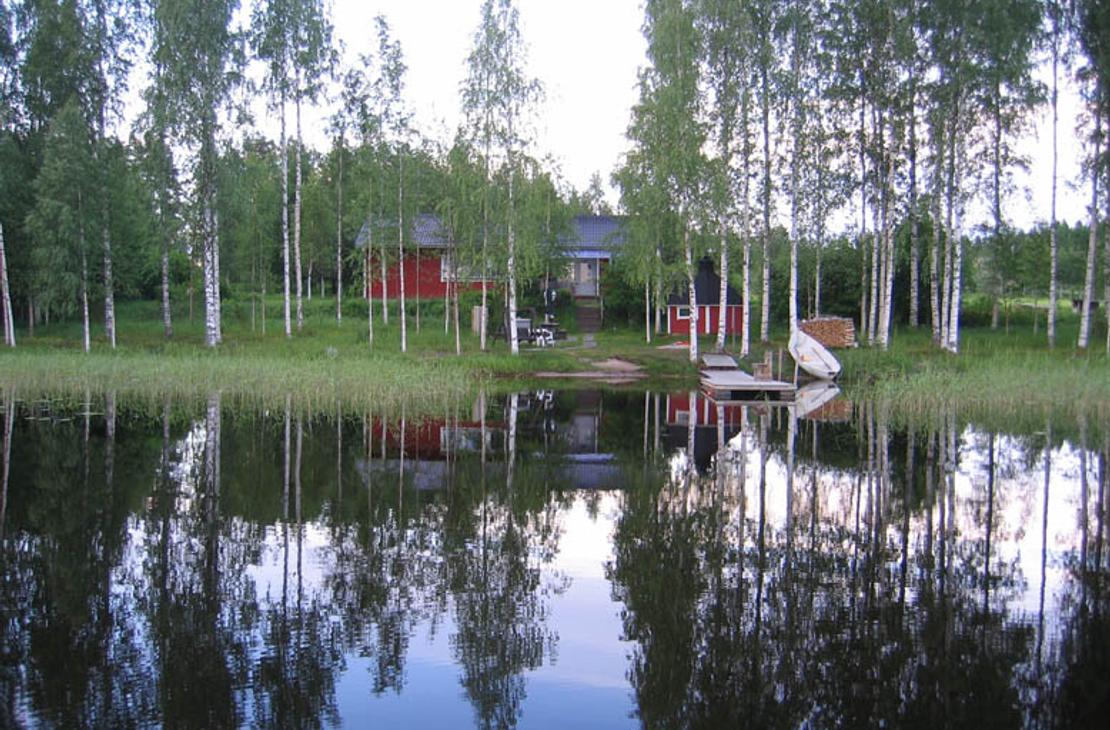 Kuva esittää Villa Blancan / LillaVilla Villan rantamaisemaa järveltä päin. Tyyni vesi etualalla heijastaa kirkkaasti taustalla olevia hoikkia koivuja ja muita puita. Puiden takaa erottuu punamultainen mökki tai sauna. Rannassa on pieni laituri ja mahdollisesti valkoinen vene tai kanootti. Maisema on rauhallinen ja kesäinen.